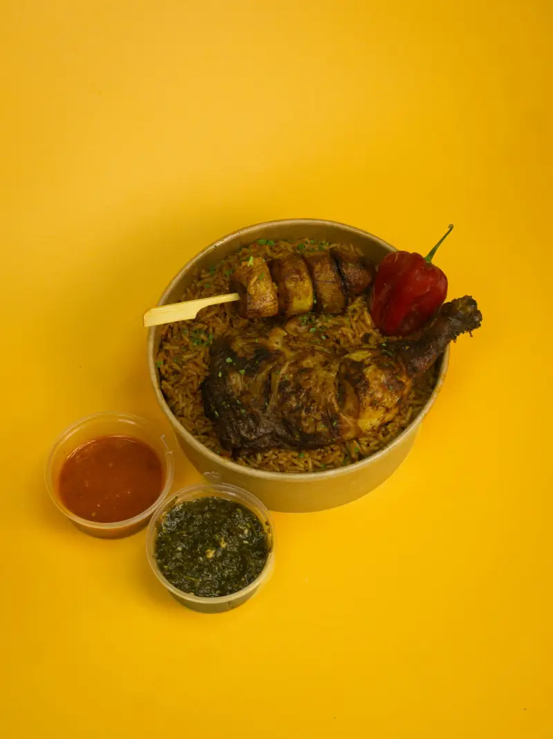 Jollof rice bowl topped with a grilled chicken leg, plantain skewer and a scotch bonnet, with two dipping pots alongside