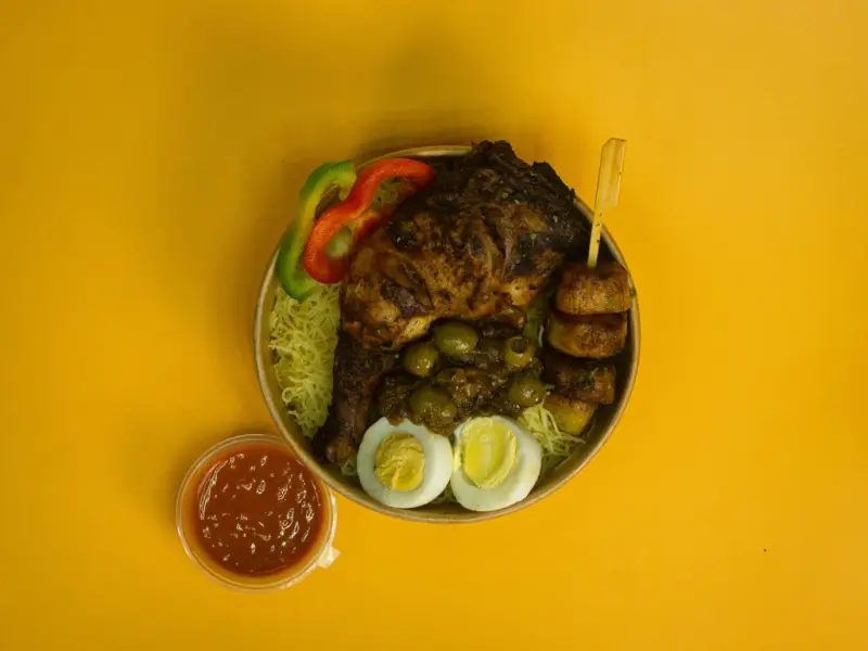 Overhead bowl of grilled chicken with olives, boiled egg, peppers and shredded salad, a pot of chilli sauce beside it