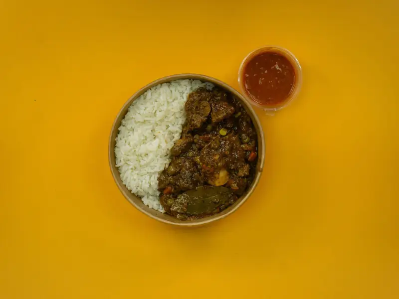 Overhead bowl of white rice with a chunky meat stew, a pot of chilli sauce set to one side