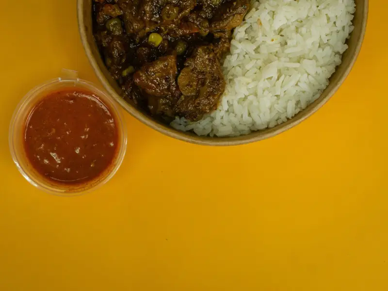 Tightly cropped corner shot of a rice and stew bowl with a pot of chilli sauce