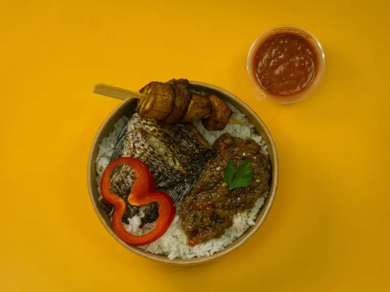 Whole grilled fish over rice with pepper sauce, a plantain skewer and a pot of chilli sauce