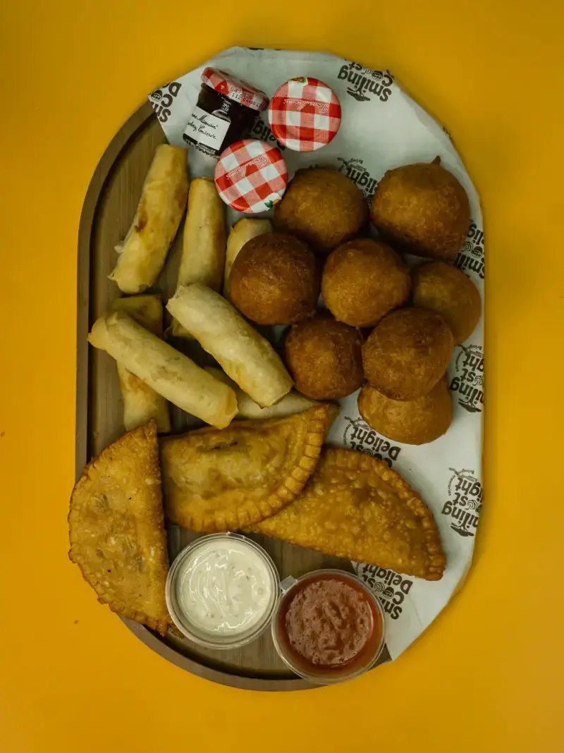 Overhead wooden board of fried dough balls, spring rolls and pastries with dipping pots