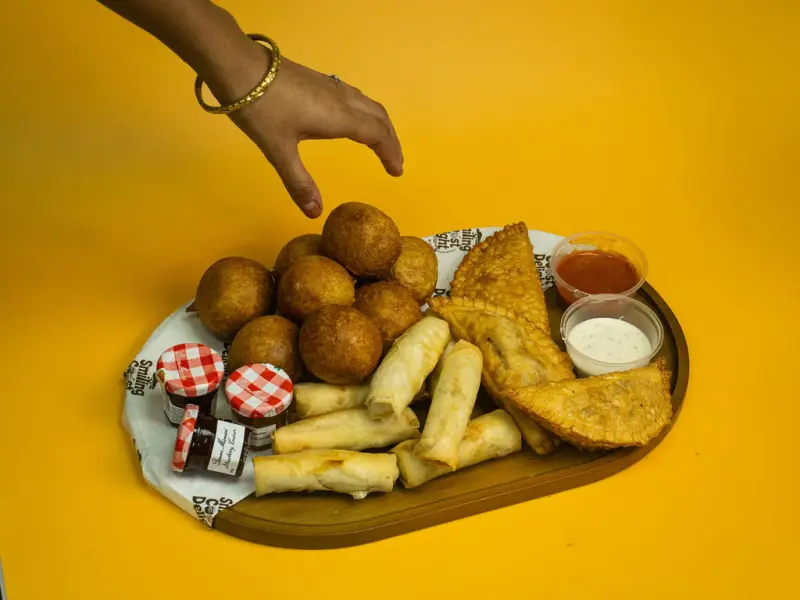 A hand reaching towards a wooden sharing board of fried dough balls, spring rolls and pastries