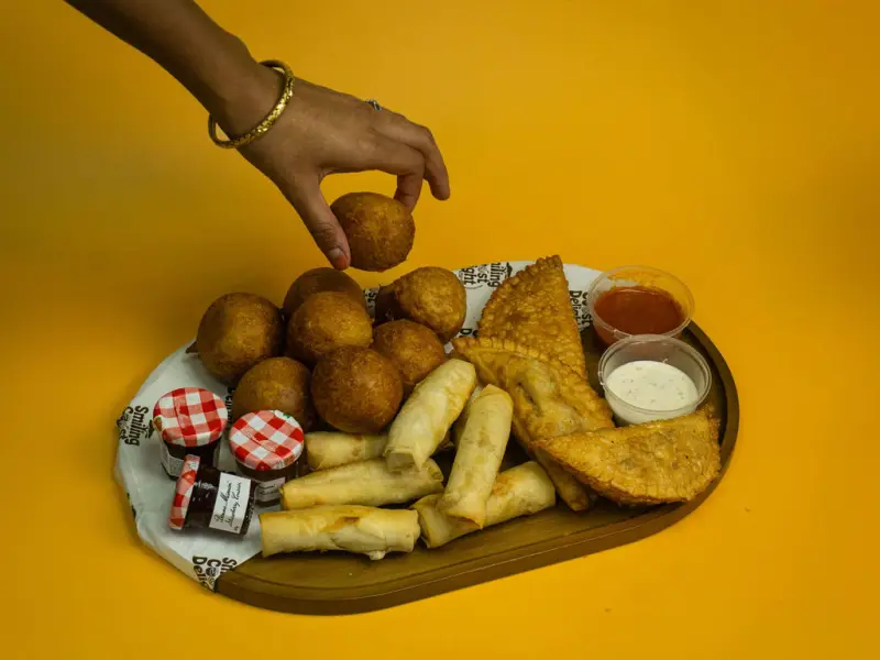 A hand lifting a fried dough ball from a wooden sharing board of snacks and dips