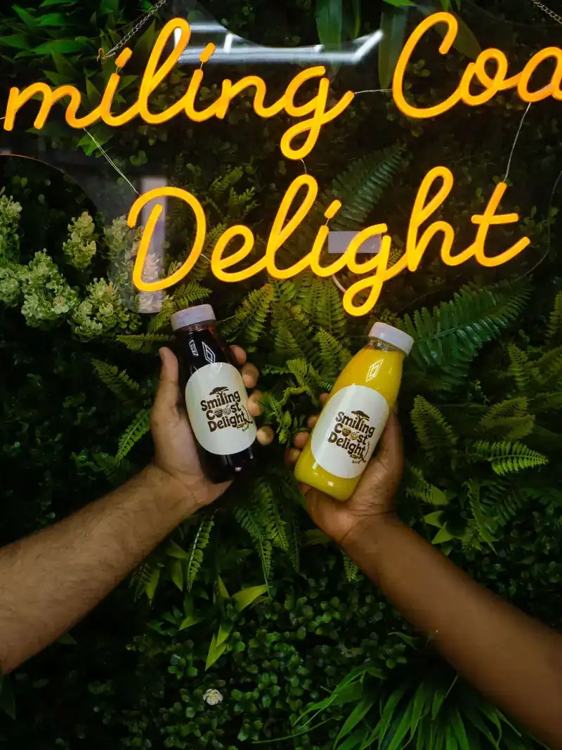 Two hands holding bottled drinks in front of an illuminated neon sign set among greenery